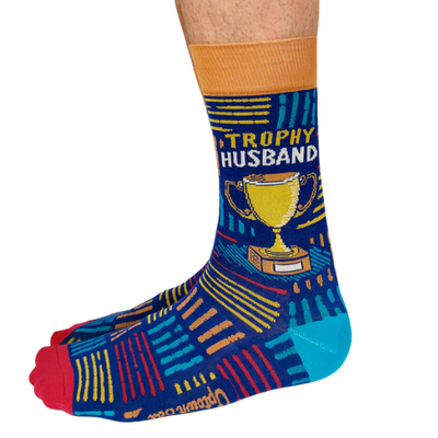 A pair of socks with a playful design featuring bold text that reads "Trophy Husband" in large, gold and white capital letters. The background is a deep, vibrant blue with small, scattered trophy icons in yellow and gold, adding a fun, humorous touch. The socks are mid-calf length and made of soft, stretchy material, perfect for a quirky gift or personal wear.